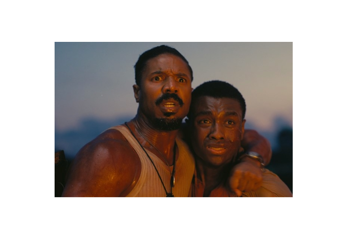 Two people standing close together outdoors during dusk, with one person’s arm around the other in a supportive gesture. Both are wearing sleeveless tops, and their skin appears glistening as if from sweat or water. The background shows a soft, blurred horizon with muted blue and orange tones, suggesting evening light. The image conveys a sense of physical exertion or intensity in a natural setting.