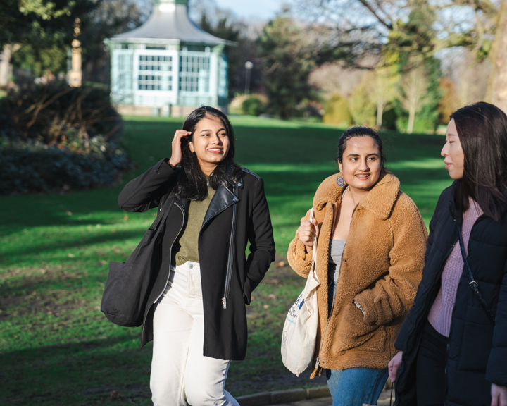 Three people walking together along a paved path in a park with green grass and trees. They are dressed in warm, casual clothing, including coats and jackets, and carrying bags. In the background, there is a light blue gazebo surrounded by greenery, and the scene is lit by natural daylight.