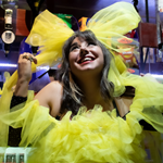 A woman in a bright yellow dress, and a huge yellow bow on her head, is smiling and looking up towards the ceiling. Behind her is a row of optics.