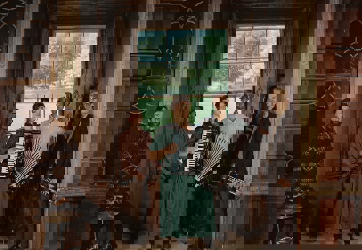  group of five musicians poses inside an ornate, old wooden interior with arched detailing and a large window looking out onto greenery. The musicians stand and sit with their instruments, including an accordion, a drum, a guitar, a double bass, and a trombone.