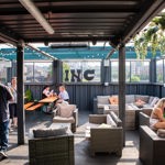 INC Sheffield - Outdoor rooftop bar with a covered seating area featuring wicker sofas with cushions, wooden tables, and string lights overhead. Green plants hang from the ceiling, and the space offers views of city buildings through mesh fencing. Several people are standing and sitting, enjoying drinks and conversation.
