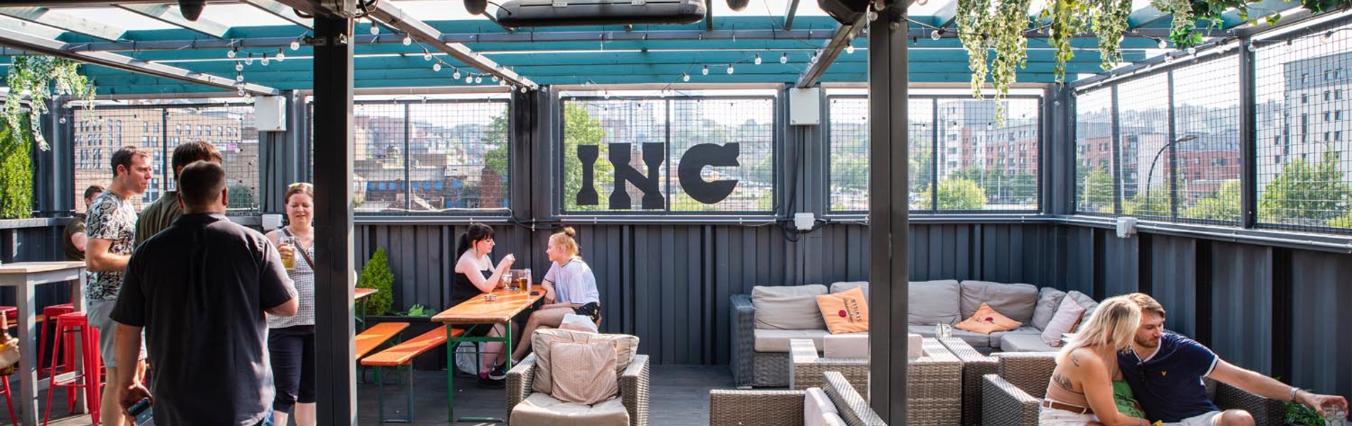 INC Sheffield - Outdoor rooftop bar with a covered seating area featuring wicker sofas with cushions, wooden tables, and string lights overhead. Green plants hang from the ceiling, and the space offers views of city buildings through mesh fencing. Several people are standing and sitting, enjoying drinks and conversation.