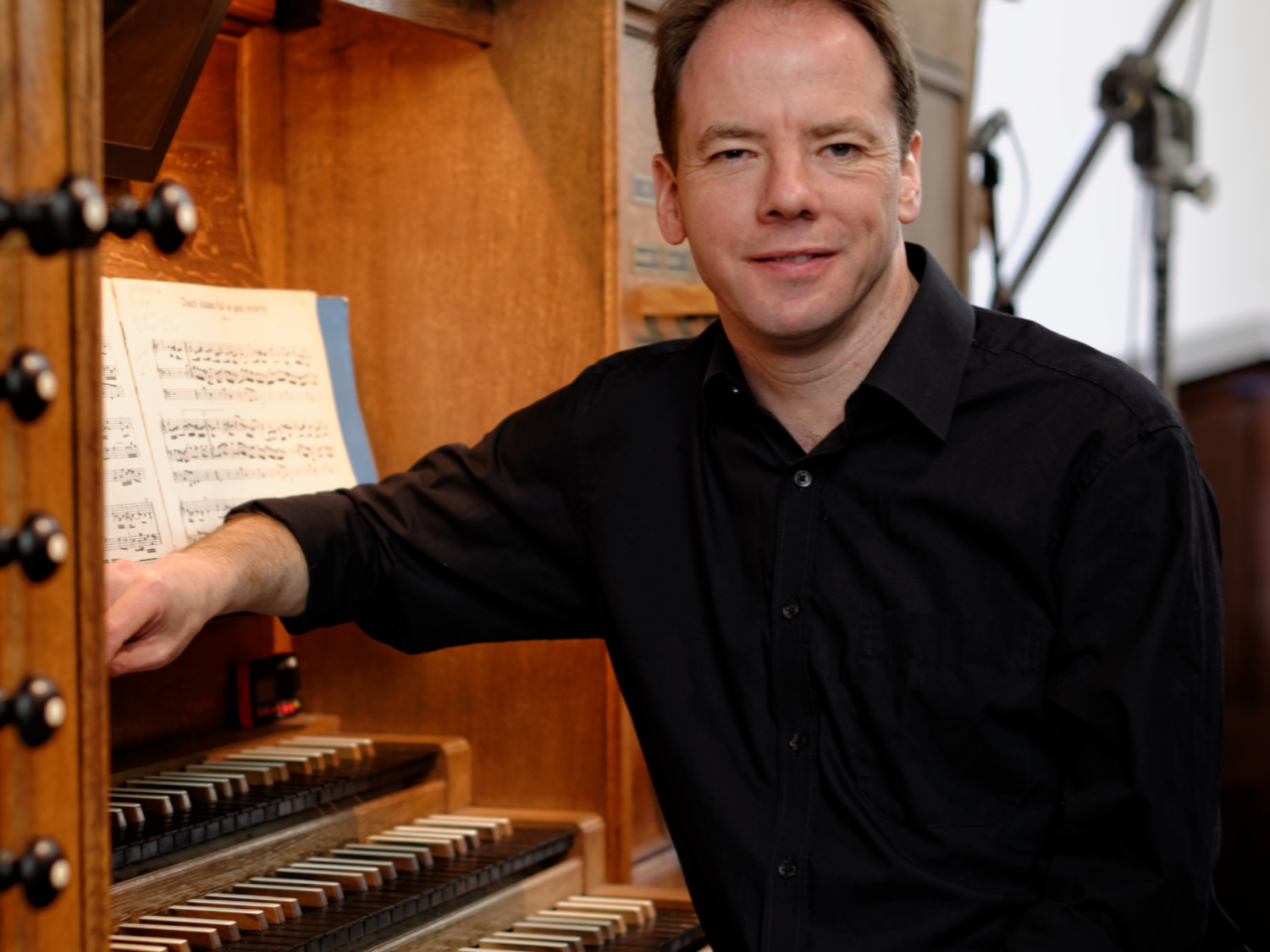 A person sits at a large pipe organ console, resting one arm beside multiple rows of keys and numerous stops. Sheet music is open above the keyboards, and the wooden organ structure surrounds them. Microphones and cables are visible in the background, suggesting a performance or recording setting.