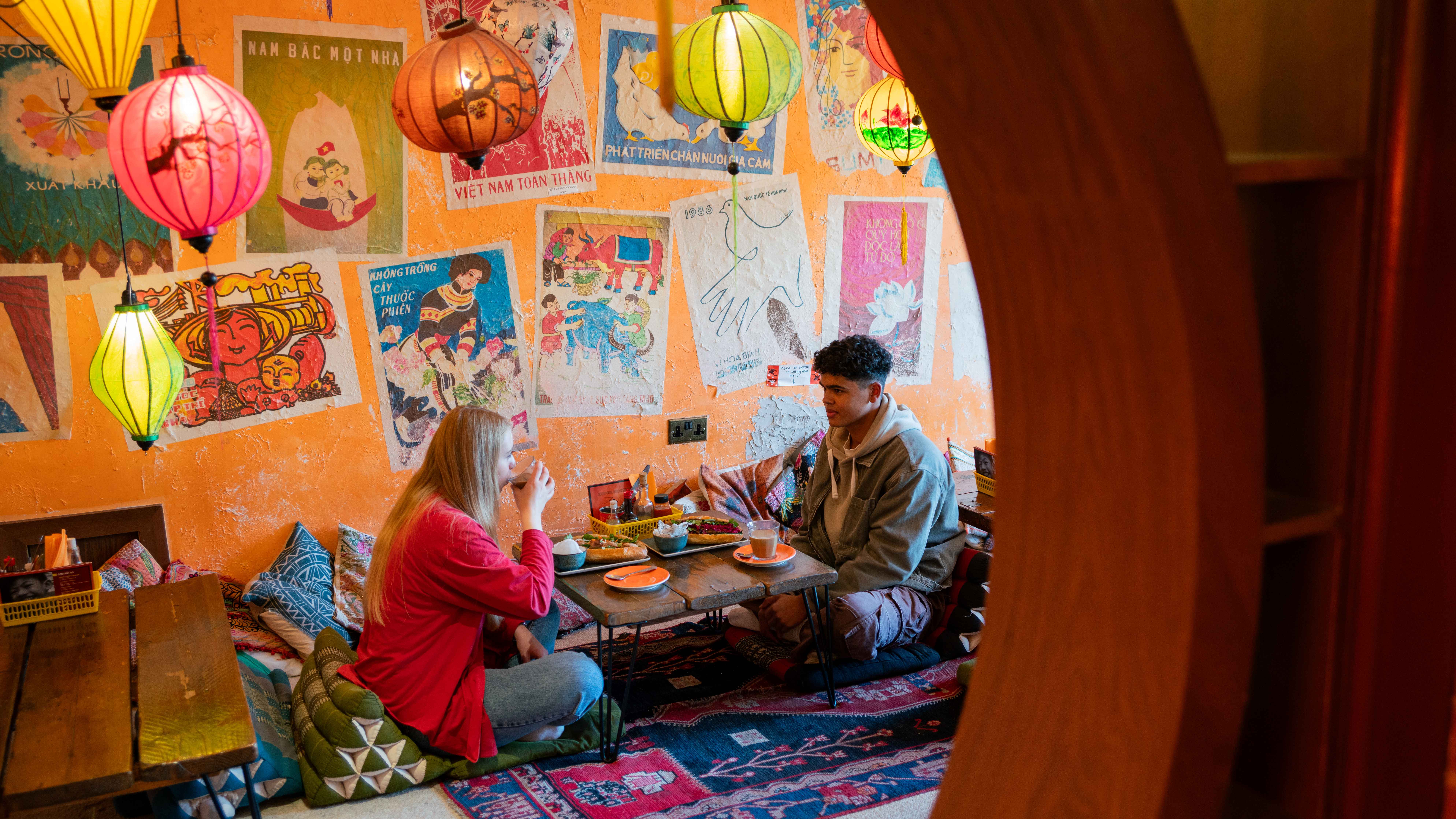 A cozy café interior with vibrant orange walls decorated with colorful Vietnamese posters and artwork. Several hanging lanterns in red, yellow, and green provide warm lighting. Two people are seated on floor cushions at a low wooden table, enjoying food and drinks. The table holds plates and cups, and the floor is covered with patterned rugs, creating a relaxed, intimate atmosphere.