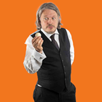 A picture of the performer, standing front of an orange background. The performer is holding a small white ball, and staring at it as though he is scared of it. He has one arm behind his back