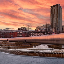 Urban plaza with cascading water features and fountains in front of a long reflective wall. Modern high-rise buildings and older brick structures are visible in the background, along with construction cranes. The sky is filled with dramatic orange and pink clouds during sunset, creating a vibrant backdrop.