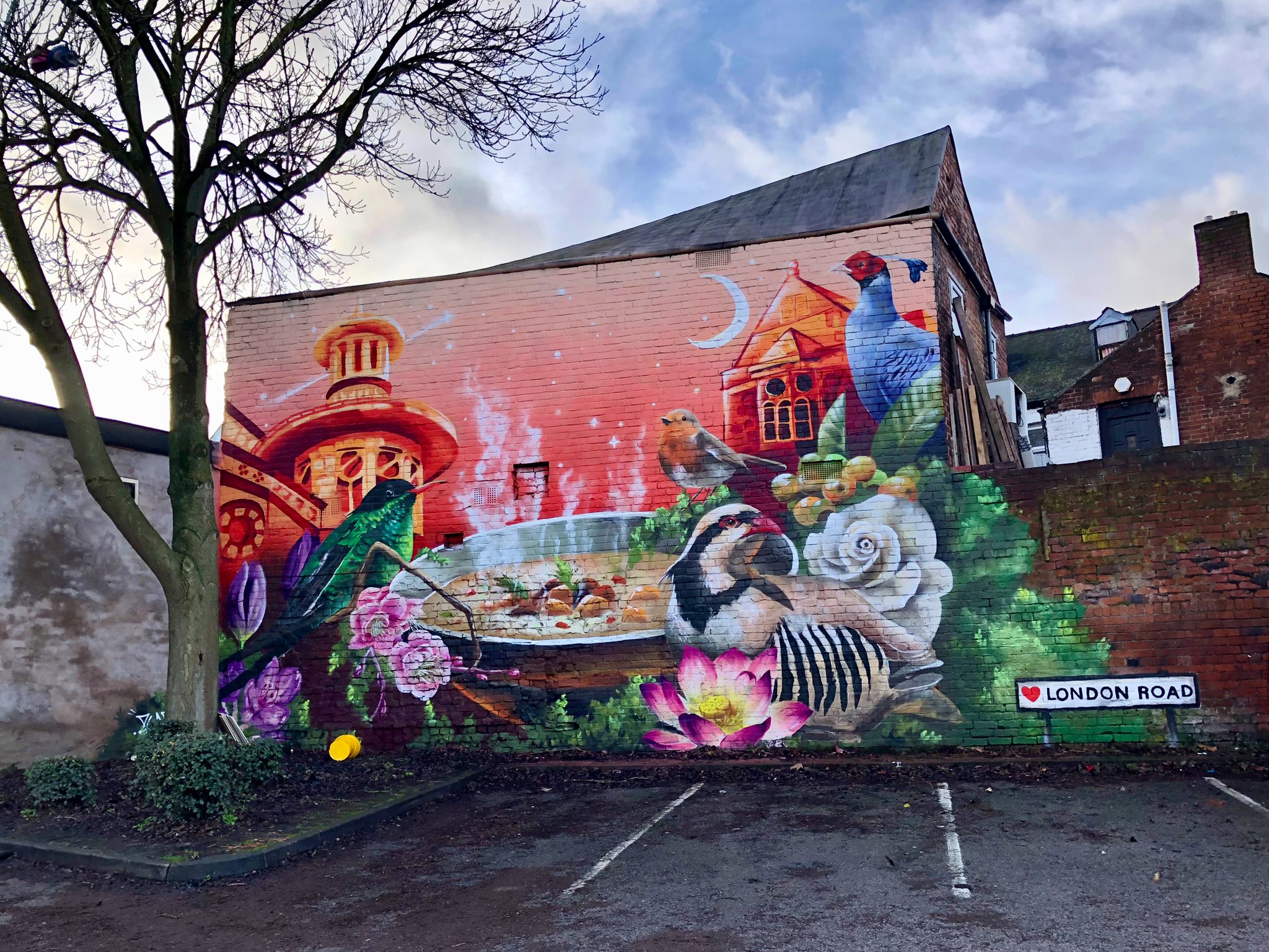 A vibrant mural painted on the side of a brick building along London Road. The artwork features a colorful nature scene with birds, flowers, and greenery, including a pheasant and a partridge near a bowl of food. Architectural elements such as domed towers and rooftops appear in the background against a red and orange sky with a crescent moon. A tree partially frames the mural on the left, and a street sign reading “♥ London Road” is visible at the bottom right.