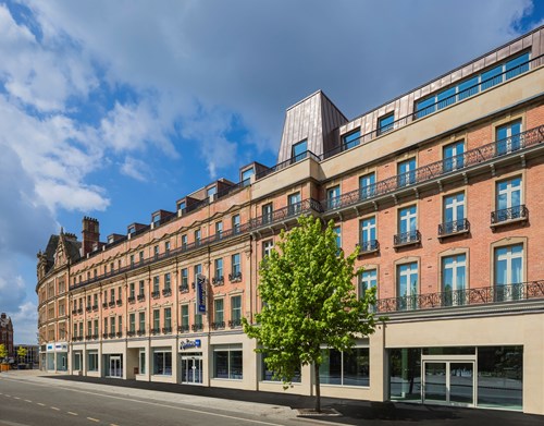 CGI-rendering of a Street view of a large, multi-story building with a red-brick façade and beige stone accents, featuring rows of tall windows with black wrought-iron balconies. The ground floor has several glass storefronts, including a visible sign for ‘Travelodge.’ A single green tree stands on the sidewalk in front of the building, and the sky above is partly cloudy with patches of blue.