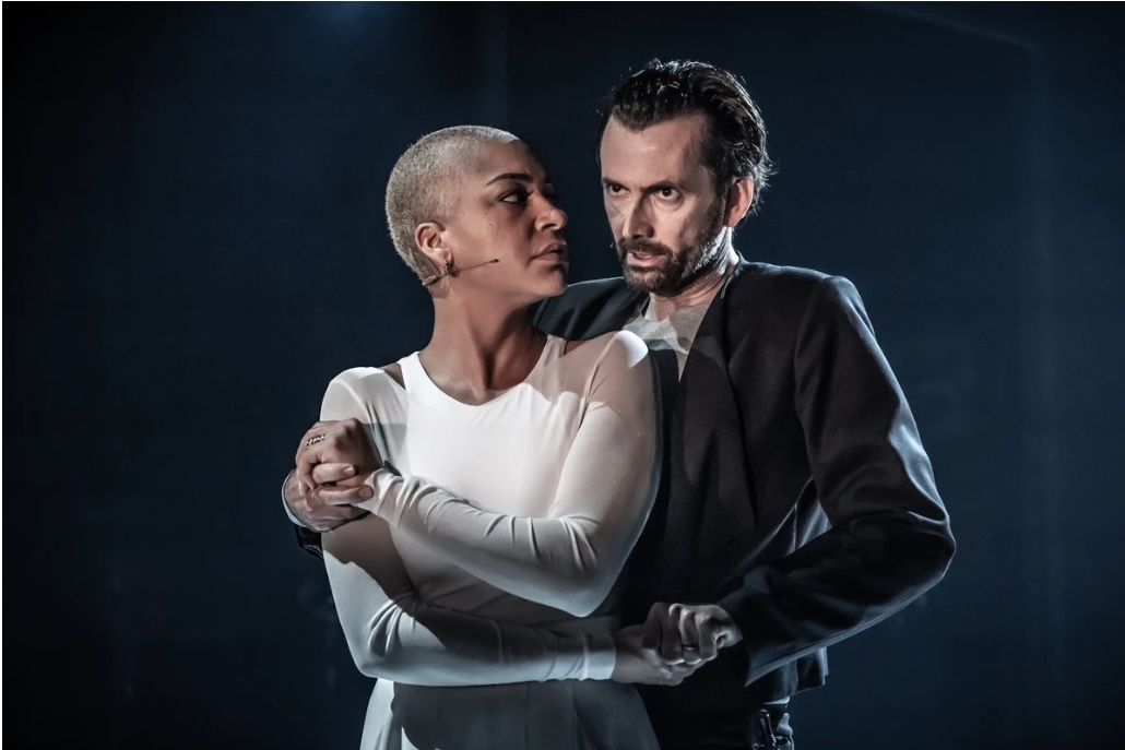 Two performers on stage in an intimate pose, with one standing behind the other and holding their arms. The person in front wears a long-sleeved white outfit, while the person behind is dressed in a dark jacket. The scene is dramatically lit with a cool, moody atmosphere, highlighting the contrast between the light and dark clothing against a dim background.