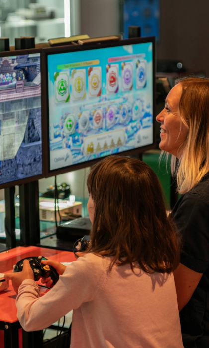 A person with a child sat next to them are playing a video game together. In the background are displays of video games from the history of the video game industry.