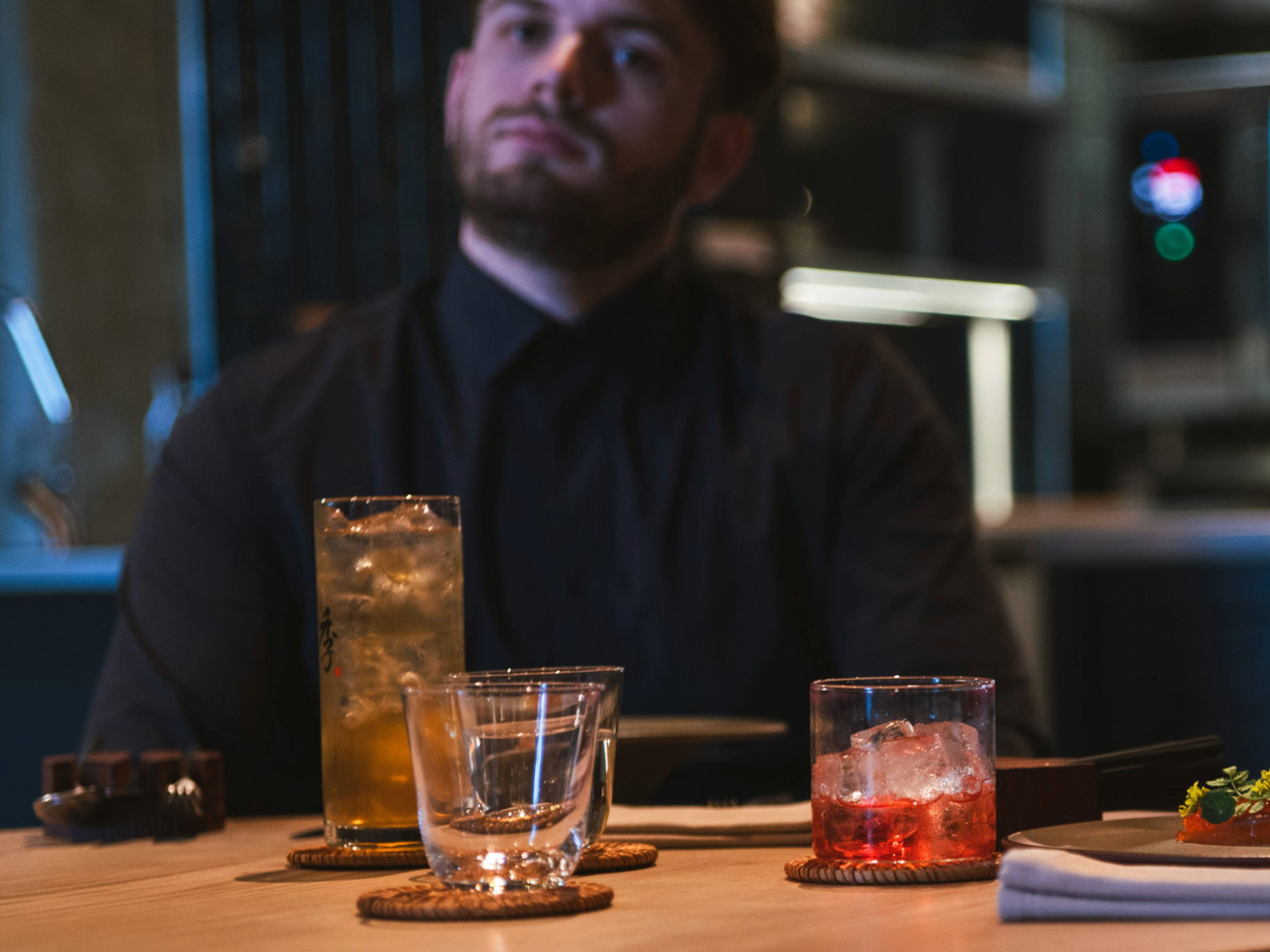 A wooden dining table in a modern restaurant setting with drinks placed on coasters. There is a tall glass of iced drink, a short glass with a pink cocktail and ice, and an empty tumbler. A folded napkin and cutlery are also visible, along with a small dish of food at the edge of the frame. The background shows a softly lit interior with sleek, contemporary design elements.