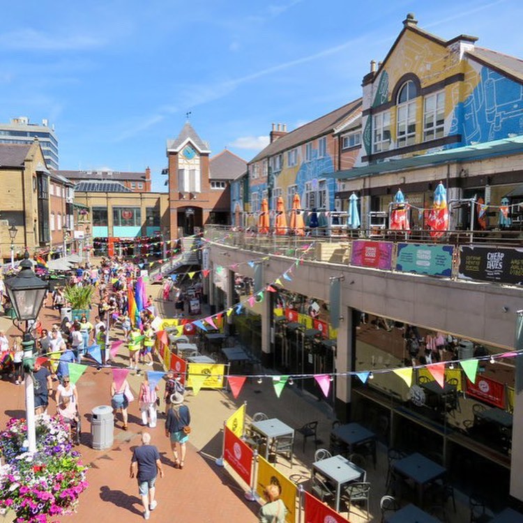 Orchard Square in the sunshine decorated with bunting. There are lots of people walking about.