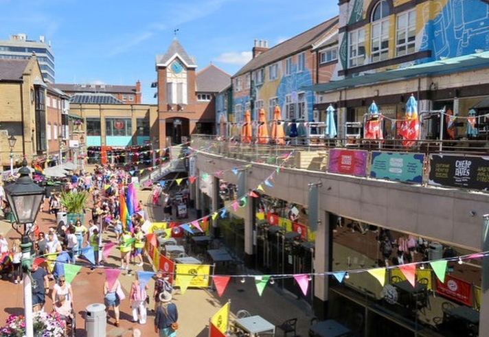 Orchard Square in the sunshine decorated with bunting. There are lots of people walking about.