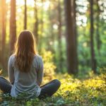 A person sits cross‑legged on the forest floor facing away from the viewer, surrounded by tall trees and lush green vegetation. Soft golden sunlight filters through the trees, illuminating patches of grass and creating a warm, tranquil atmosphere. The scene conveys a peaceful moment of stillness in nature, with the person seated at the center of the sunlit woodland.