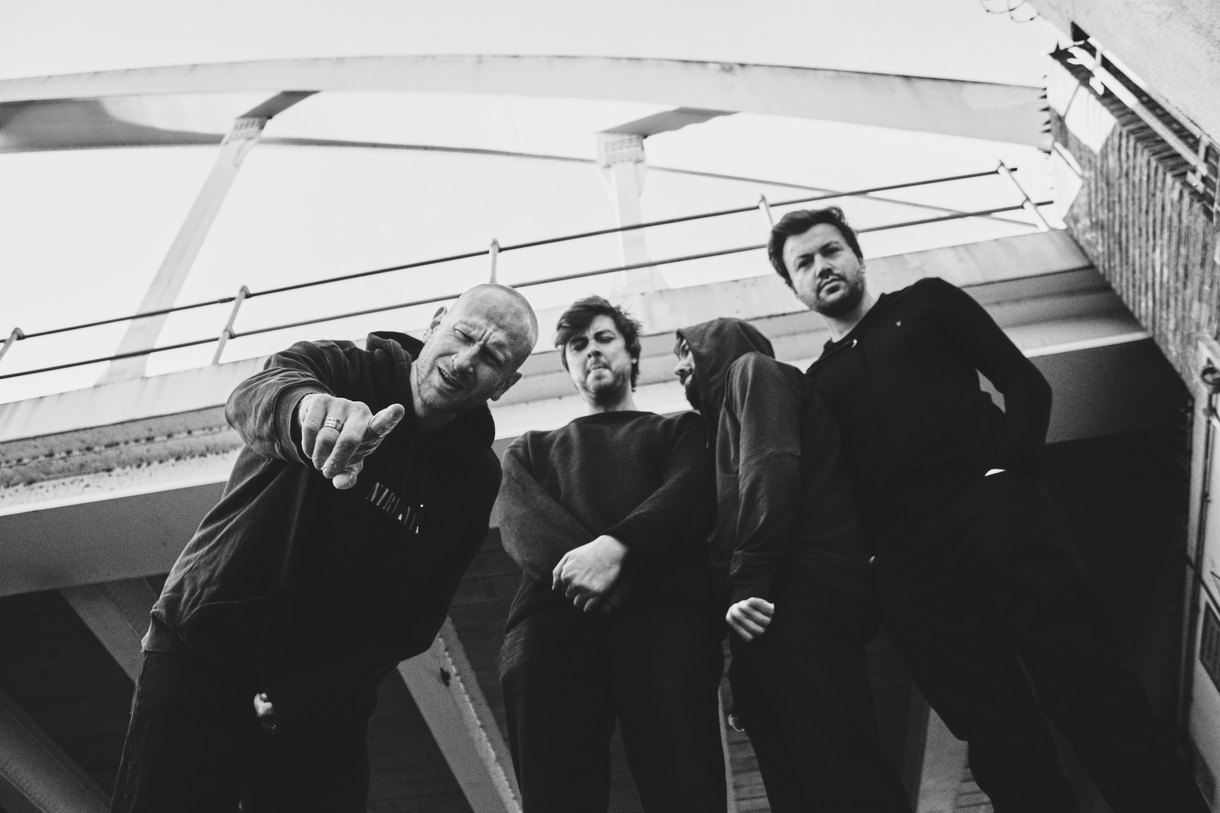 A black‑and‑white, low‑angle shot of four people standing beneath a modern bridge structure. They wear dark, casual clothing, with one person leaning forward and gesturing towards the camera. The group stands close together, framed by the sweeping lines of the bridge above.