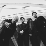 A black‑and‑white, low‑angle shot of four people standing beneath a modern bridge structure. They wear dark, casual clothing, with one person leaning forward and gesturing towards the camera. The group stands close together, framed by the sweeping lines of the bridge above.