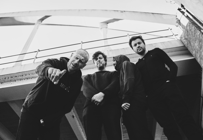 A black‑and‑white, low‑angle shot of four people standing beneath a modern bridge structure. They wear dark, casual clothing, with one person leaning forward and gesturing towards the camera. The group stands close together, framed by the sweeping lines of the bridge above.