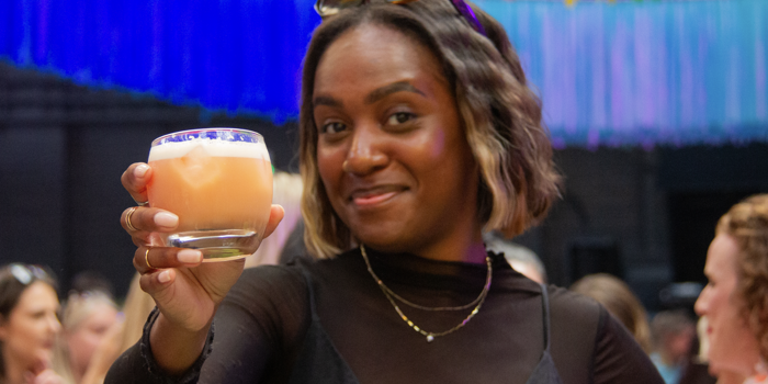 A person at a lively event holds up a glass of orange‑coloured drink as if making a toast. They wear dark, stylish clothing with layered necklaces and sunglasses resting on their head. Behind them, colourful hanging decorations in bright rainbow shades fill the space, with a crowd of people gathered in the background.