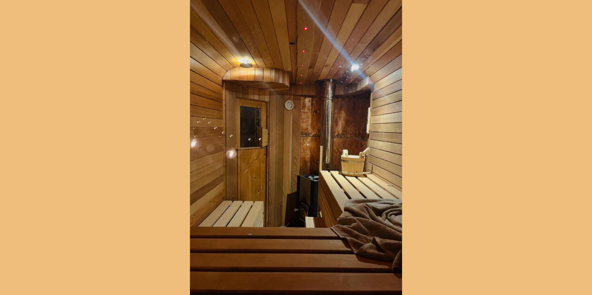 A warm, wood‑panelled sauna interior with benches on both sides and soft lighting reflecting off the curved ceiling. A towel rests on one of the benches, and a small stove with stones sits near the back. A wooden bucket and ladle are placed on a shelf beside the stove, creating a calm, inviting atmosphere.