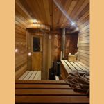 A warm, wood‑panelled sauna interior with benches on both sides and soft lighting reflecting off the curved ceiling. A towel rests on one of the benches, and a small stove with stones sits near the back. A wooden bucket and ladle are placed on a shelf beside the stove, creating a calm, inviting atmosphere.