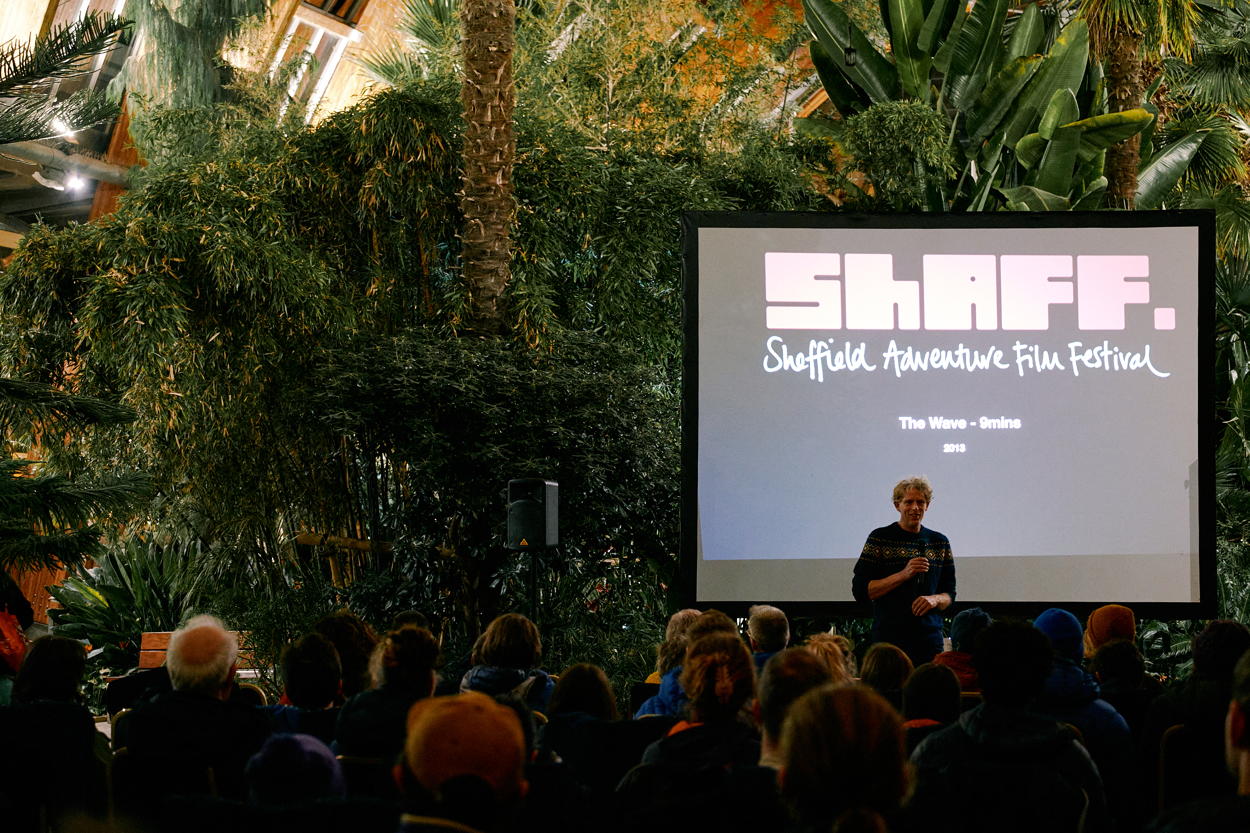 Man standing in front of a black screen with the Sheffield Adventure Film Festival (SHAFF) logo, talking to a crowd of people in Sheffield Winter Garden