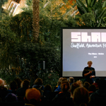 Man standing in front of a black screen with the Sheffield Adventure Film Festival (SHAFF) logo, talking to a crowd of people in Sheffield Winter Garden