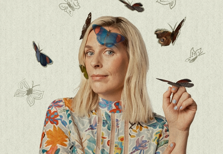 A portrait photograph of Sarah Pascoe. She is wearing a floral dress and is smiling wryly at the camera. There are butterflies on her face and one on her hand. There are also butterflies drawn on the background.