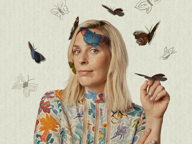 A portrait photograph of Sarah Pascoe. She is wearing a floral dress and is smiling wryly at the camera. There are butterflies on her face and one on her hand. There are also butterflies drawn on the background.