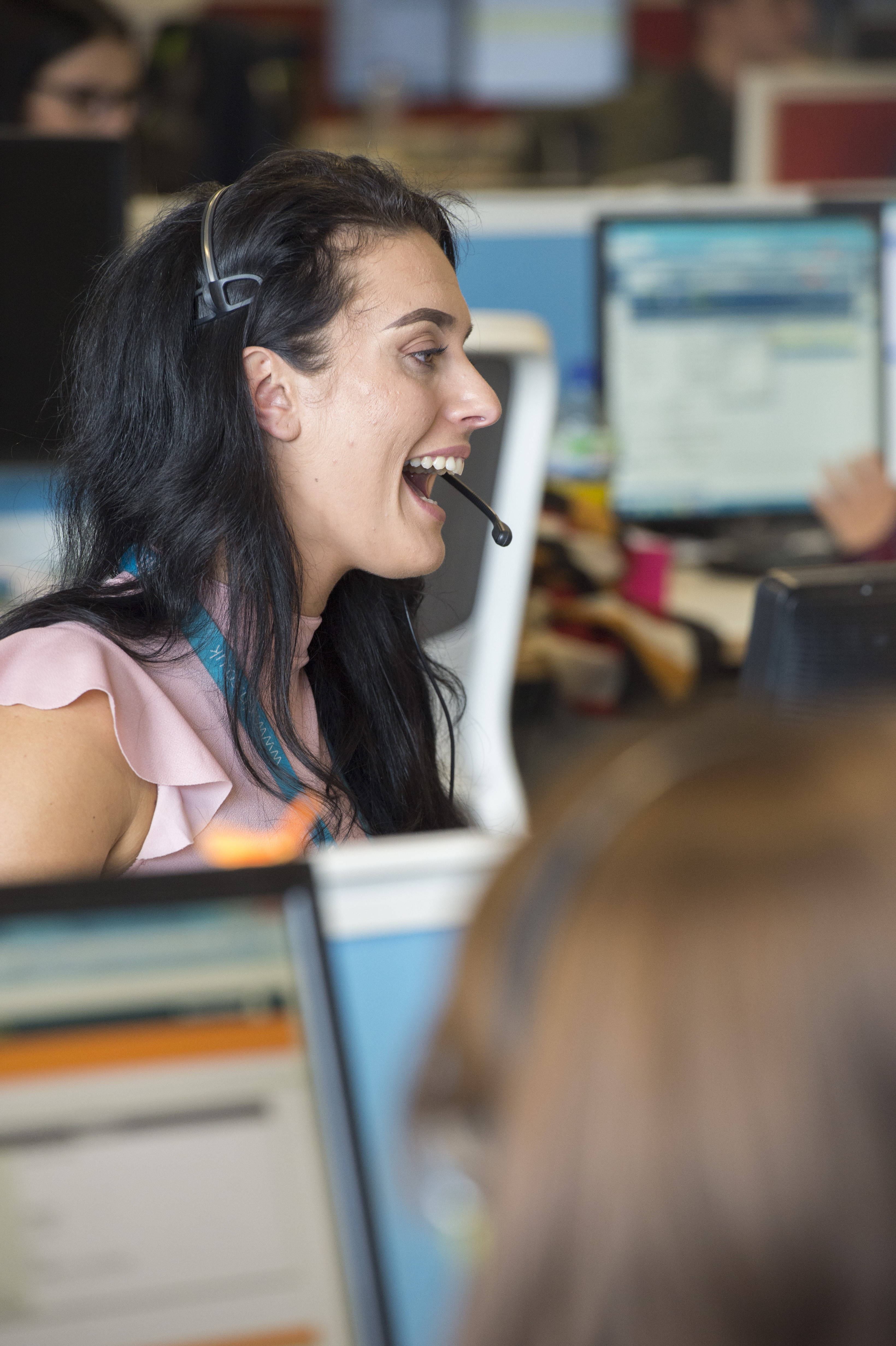 A young woman in a call centre is wearing a headset and is talking animatedly and smiling.