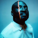 Joshua Idehen, looking upwards, in subdued blue lighting.