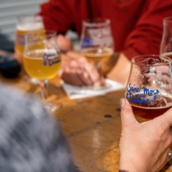 Close-up of several people sitting around a wooden table with multiple glasses of craft beer. One hand in the foreground holds a glass labeled ‘Saint Mars of the Desert.’ Other glasses with similar labels and varying shades of beer are visible, along with a notepad and pen on the table.