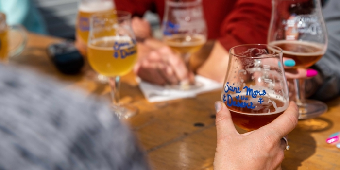 Close-up of several people sitting around a wooden table with multiple glasses of craft beer. One hand in the foreground holds a glass labeled ‘Saint Mars of the Desert.’ Other glasses with similar labels and varying shades of beer are visible, along with a notepad and pen on the table.