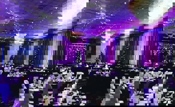 Large banquet hall decorated for an evening event with round tables covered in black cloths and white chair covers. Each table is set with white napkins, menus, and tea cups. The room features two large crystal chandeliers, purple uplighting along the walls, and strings of fairy lights draped across the ceiling. Christmas trees with lights are placed near the walls, and a stage with a backdrop of twinkling lights is visible at the far end.