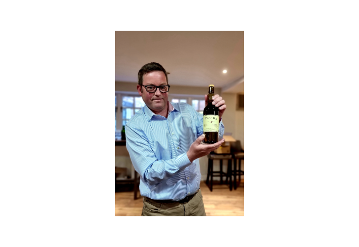 A person wearing a light blue, long-sleeved shirt is holding a bottle of Caol Ila 12-year-old single malt Scotch whisky with both hands. The bottle has a cream label with black and green text. The setting appears to be indoors, with wooden chairs and tables in the background and soft lighting from windows.