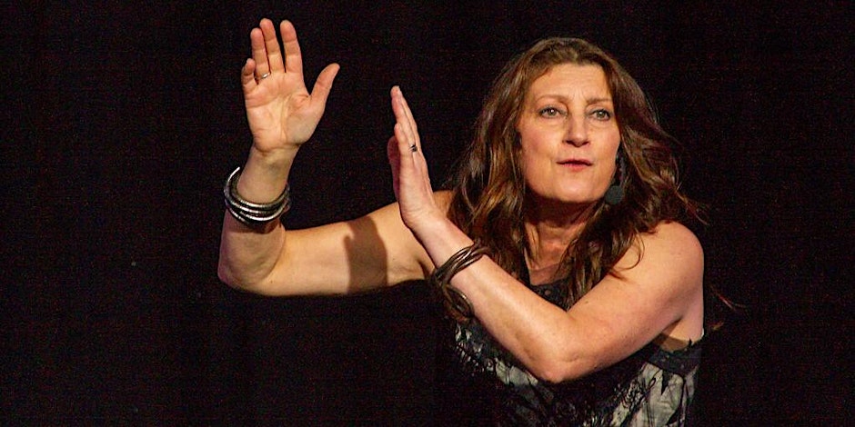 A person performing on stage with arms raised in a dynamic pose, wearing multiple bangles and a sleeveless top. The background is dark, drawing attention to the expressive movement and dramatic lighting.
