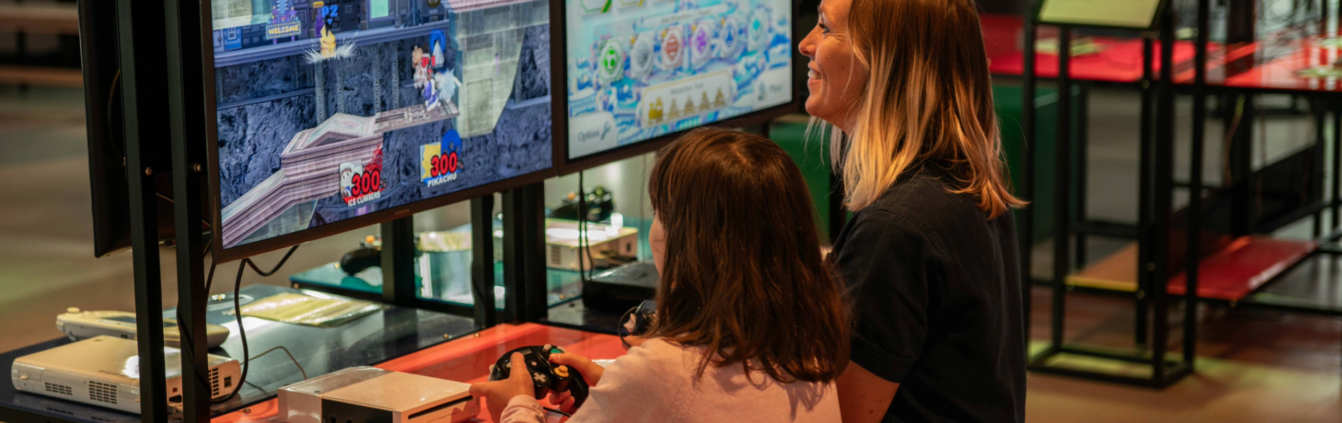 A person with a child sat next to them are playing a video game together. In the background are displays of video games from the history of the video game industry.