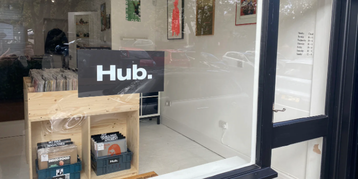 The window of a record store with a sign in the window that reads 'HUB.' . In the shop you can see wooden racks filled with vinyl records.