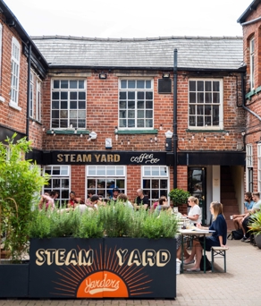 Outdoor courtyard of Steam Yard café with people sitting at tables, surrounded by red‑brick buildings and greenery.