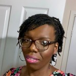 Close-up of a person wearing a floral-patterned top with pink, green, and orange flowers against a dark background. The image focuses on the upper body and hairstyle.