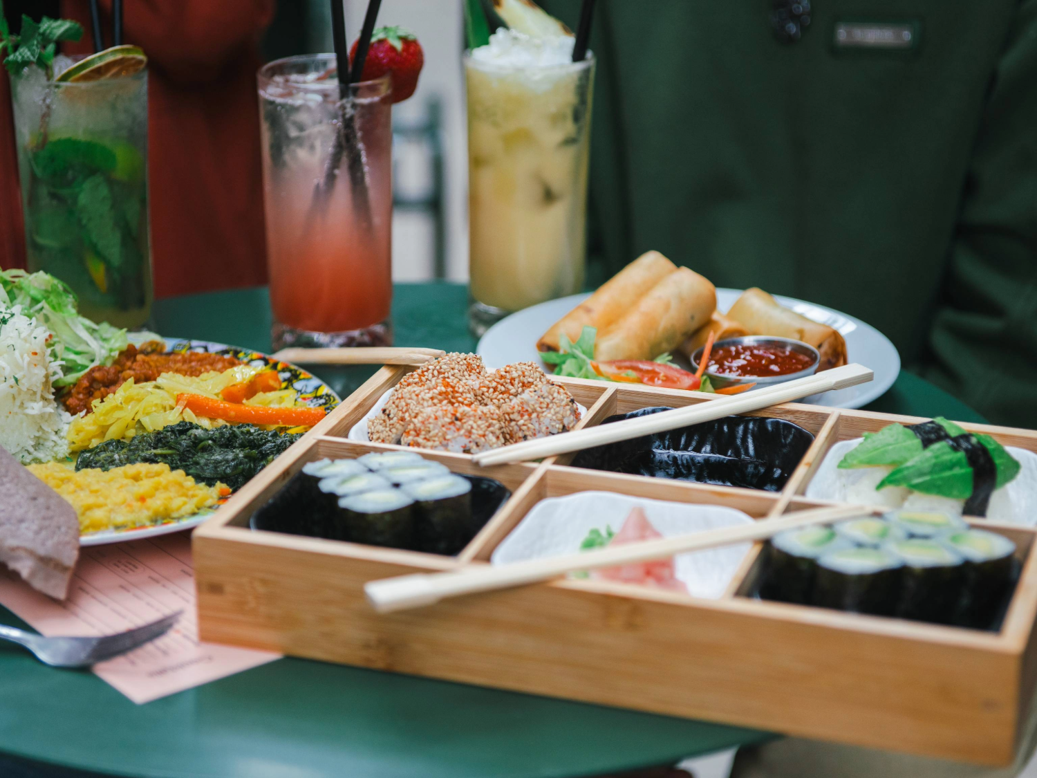 A green table filled with assorted dishes and drinks. A wooden bento-style tray holds sushi rolls, nigiri topped with avocado, sesame-coated pieces, and pickled ginger, with chopsticks resting across it. Behind the tray, a plate of golden spring rolls with dipping sauce and salad is visible, alongside a colourful platter of flatbread, shredded cabbage, and vibrant vegetable sides. Three tall cocktails garnished with fruit and herbs complete the vibrant dining scene.