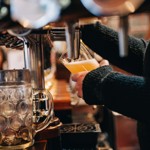 A close-up of a hand holding a glass under a beer tap as a pale, frothy beer is poured. A large empty stein sits on the bar beside it.