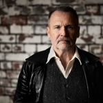 A headshot of Martin Simpson, who is stood in front of a brick wall with peeling white paint.