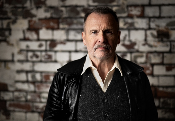 A headshot of Martin Simpson, who is stood in front of a brick wall with peeling white paint.