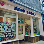 Street‑level view of the Juno Books storefront, showing a cream‑coloured façade with large windows, a sign reading ‘JUNO BOOKS’, and a colourful display in the left window featuring a handwritten message surrounded by bright paper decorations. The interior of the bookshop is visible through the right window, showing bookshelves and vibrant décor.”