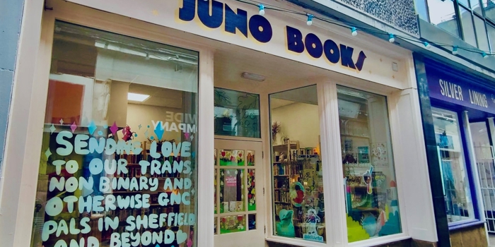 Street‑level view of the Juno Books storefront, showing a cream‑coloured façade with large windows, a sign reading ‘JUNO BOOKS’, and a colourful display in the left window featuring a handwritten message surrounded by bright paper decorations. The interior of the bookshop is visible through the right window, showing bookshelves and vibrant décor.”