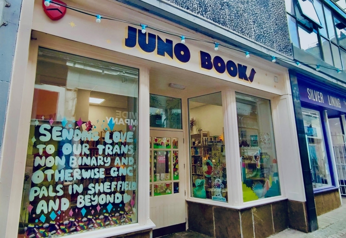 Street‑level view of the Juno Books storefront, showing a cream‑coloured façade with large windows, a sign reading ‘JUNO BOOKS’, and a colourful display in the left window featuring a handwritten message surrounded by bright paper decorations. The interior of the bookshop is visible through the right window, showing bookshelves and vibrant décor.”