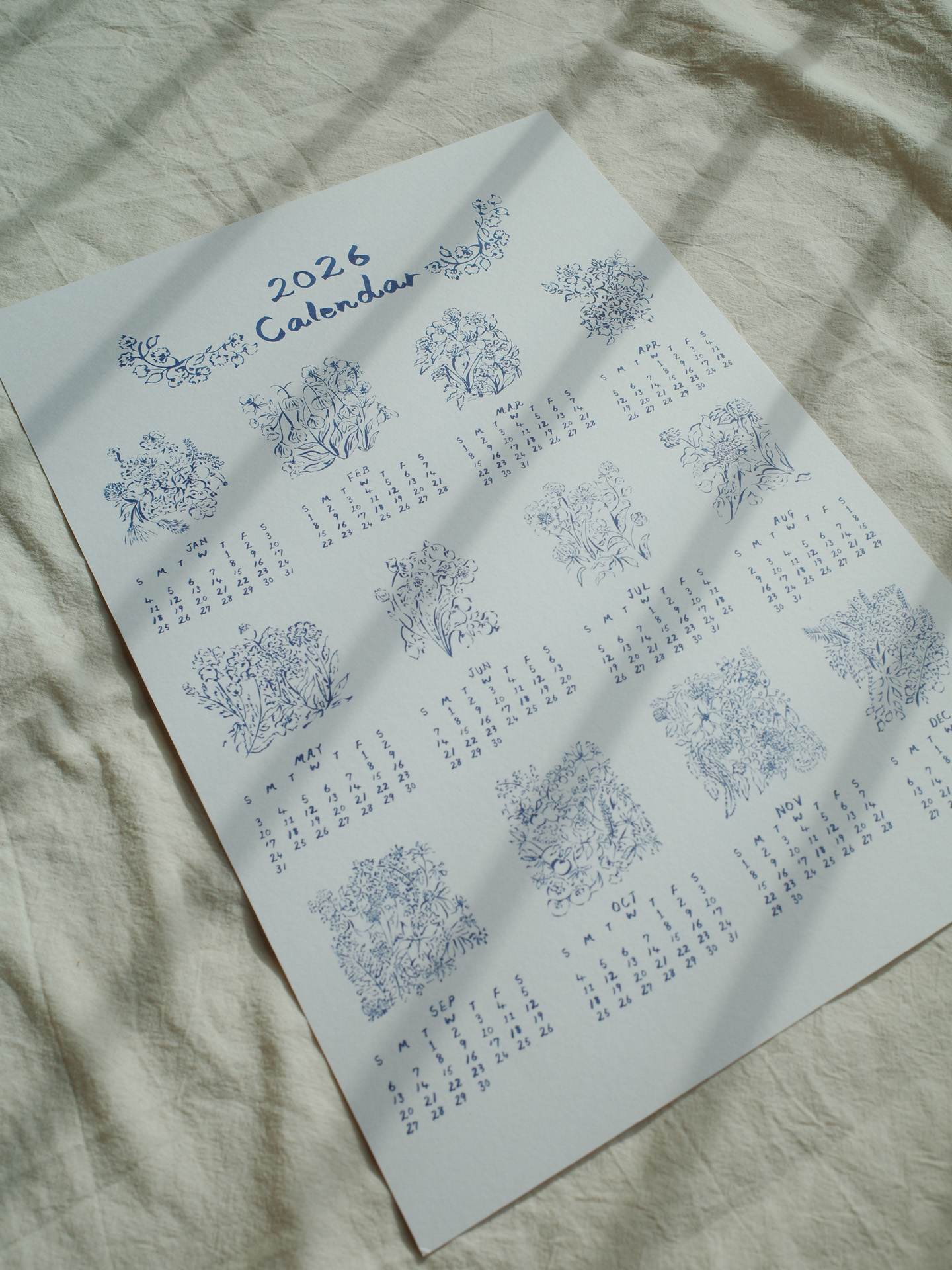 A single-page 2026 calendar printed in blue ink on white paper, featuring hand-drawn floral illustrations for each month. The calendar is laid flat on a light fabric surface with soft shadows cast across it.