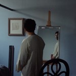 A man in a dimly lit room contemplates his reflection in a large mirror.