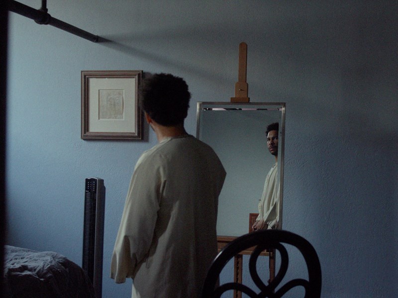 A man in a dimly lit room contemplates his reflection in a large mirror.