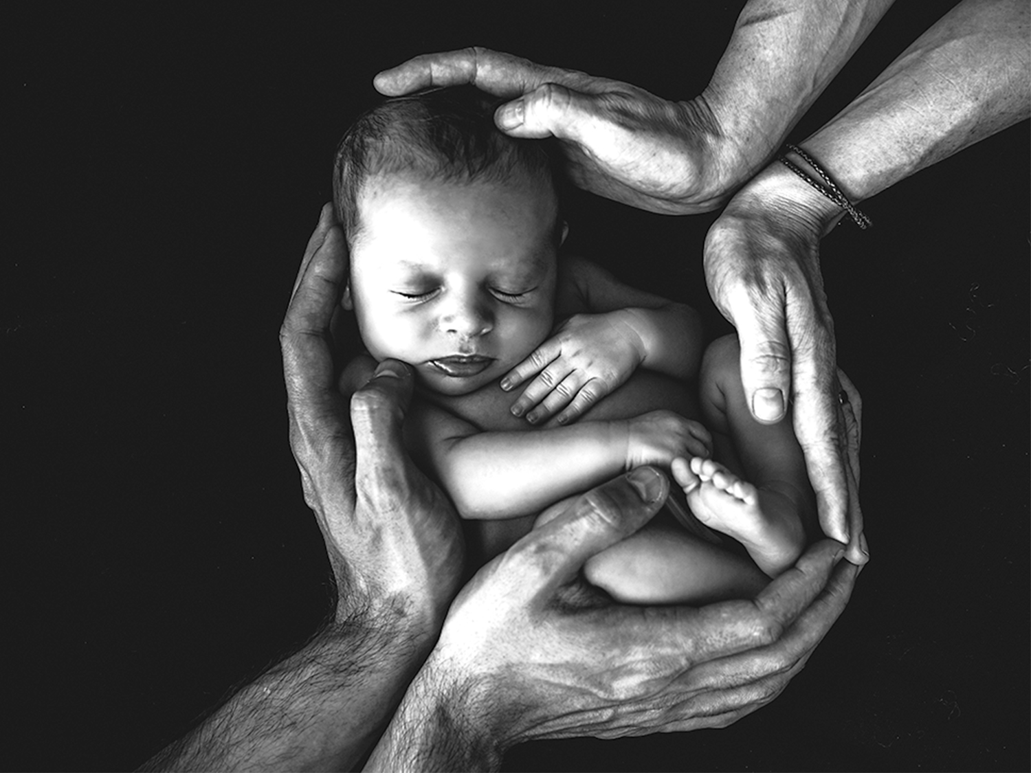 Black and white photo of a newborn baby cradled in multiple adult hands forming a protective circle around the baby’s body, set against a dark background.